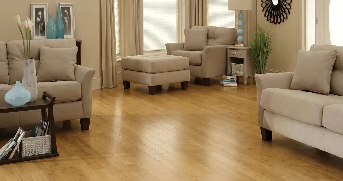 A living room with professionally installed bamboo flooring, beige sofas, a matching ottoman, a side table with a blue lamp, vases, framed pictures, and large windows with blinds and curtains in Naperville, IL
